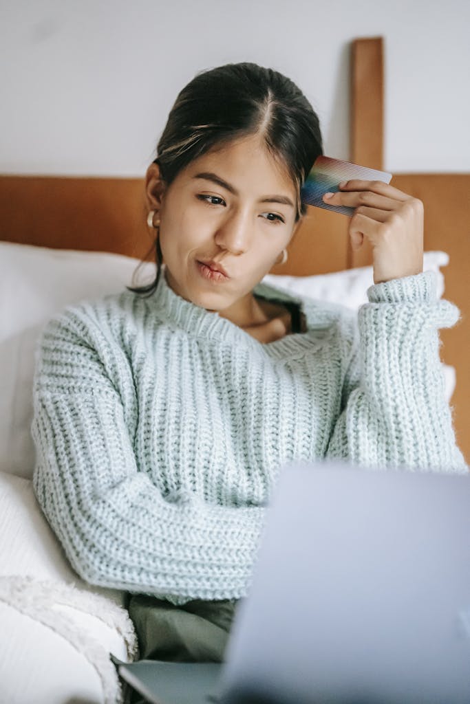 Pensive ethnic shopper with debit card and laptop on bed